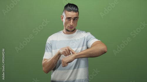 Photography Man points finger to wrist and taps bare wrist in studio with green backdrop, wearing striped tshirt and visible ring on left hand; impatience time reminder