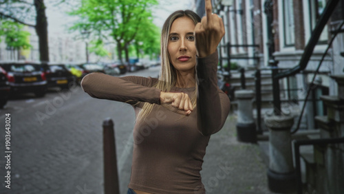 Fotografija Woman shows middle finger and clenched fist on city street, wearing brown top and looking forward; defiance empowerment