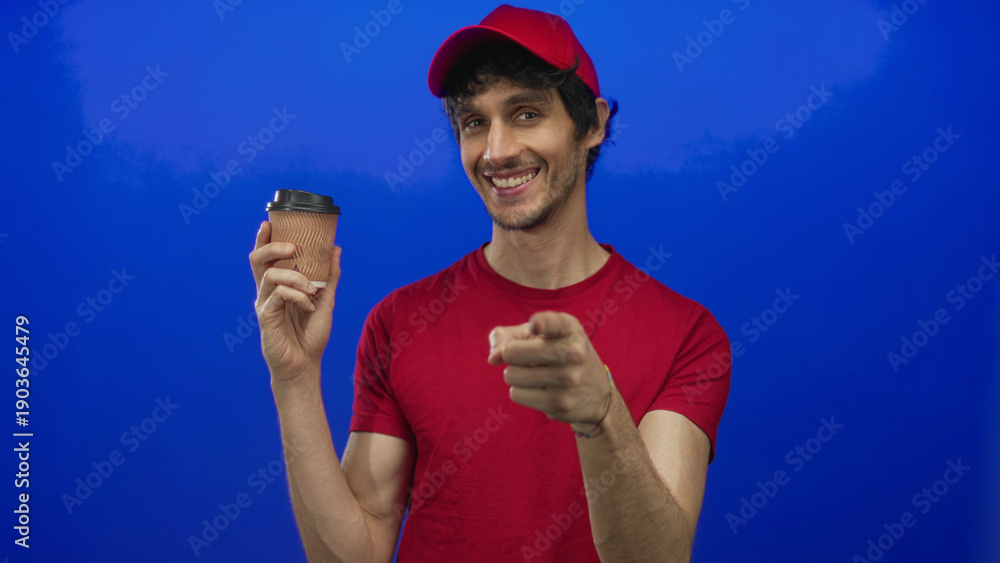 Naklejka premium Man wearing red cap and t shirt holds ribbed coffee cup and points finger toward viewer in vivid blue studio; invitation.