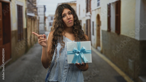 Canvas-taulu Woman holding gift box with blue ribbon in an old street, sleeveless top and curly hair, offers it with hesitant expression; reluctant offering uncertainty