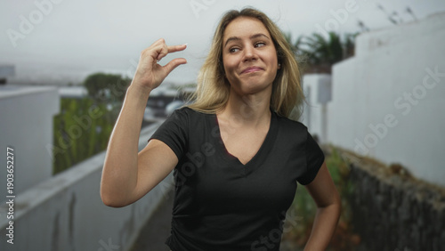 Wallpaper Mural Blonde young woman pinching fingers to show a small gap on a narrow street with white walls; playfulness confidence. Torontodigital.ca