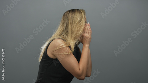 Obraz na plátně Woman covering face with hands, bare arms visible, blonde hair and black dress against gray studio wall; sadness solitude reflection