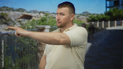 Fotografija Man points finger at camera on street walkway near metal railing and rocky bluff, wearing beige polo and trimmed beard; commanding confidence