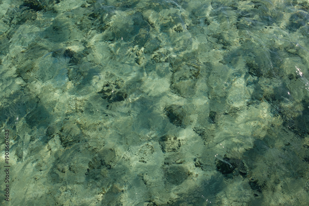 Fototapeta premium Clear turquoise water surface with visible rocks and sand beneath, creating a natural underwater scene in a tropical setting