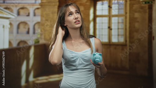 Free Download Young woman holding piggy bank with hand to neck gesture in old town building corridor; financial worry savings. - GFXLoad