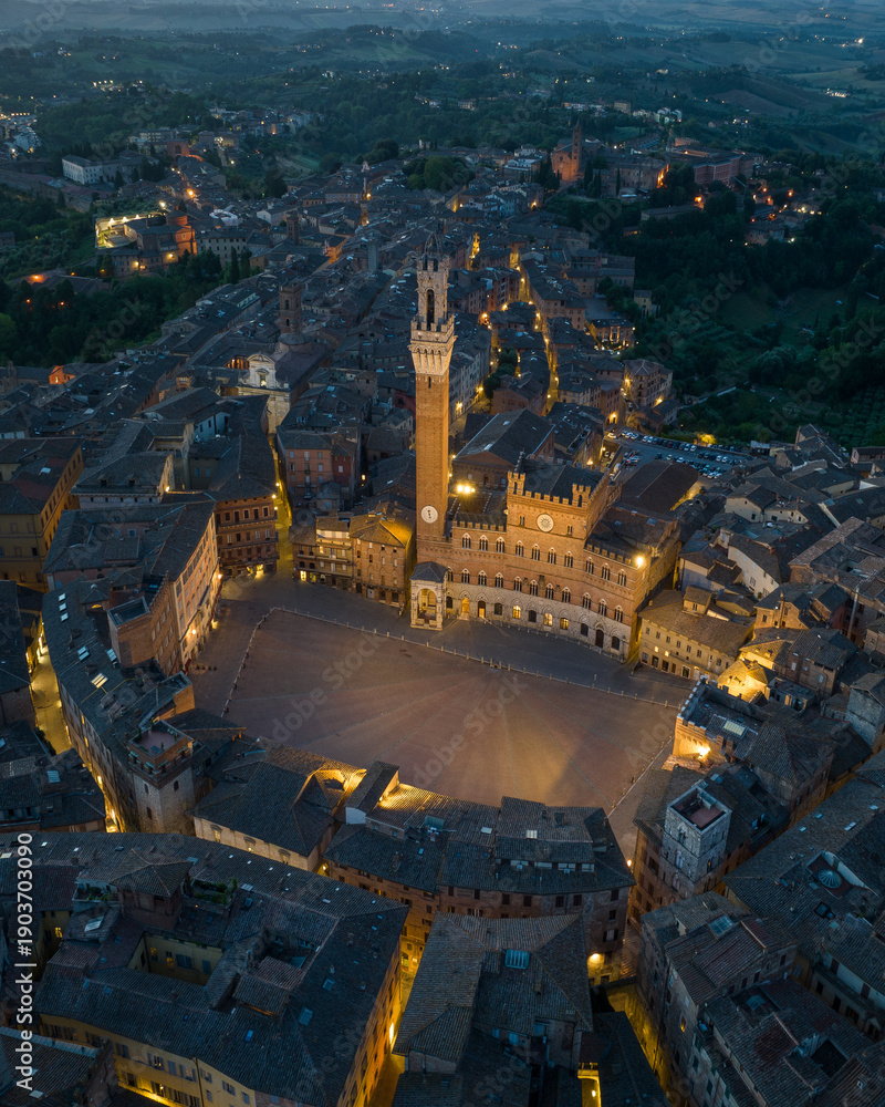 Fototapeta premium Aerial night view of Torre del Mangia rising above Piazza del Campo in Siena, Tuscany. Illuminated medieval tower and historic city center seen from above during evening hours