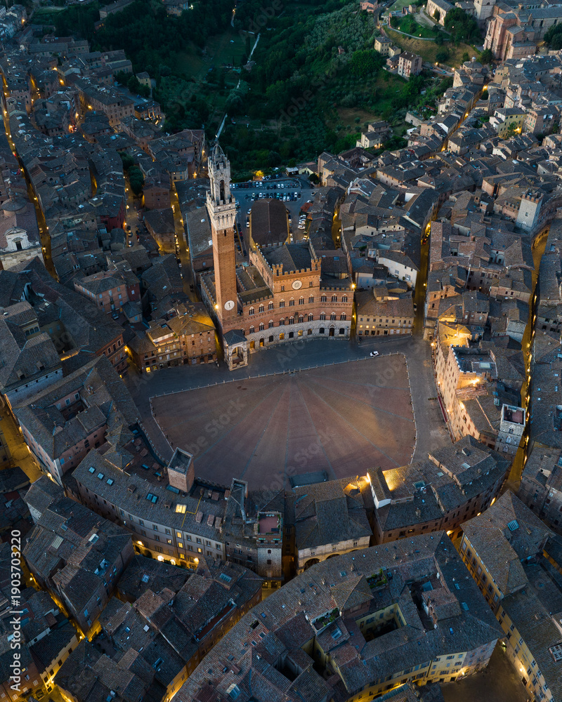 Fototapeta premium Aerial night view of Torre del Mangia rising above Piazza del Campo in Siena, Tuscany. Illuminated medieval tower and historic city center seen from above during evening hours