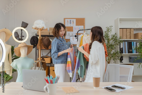 Two cheerful Asian women in a fashion studio happily taking a selfie while holding colorful clothes, enjoying creative teamwork, online selling, and modern small business lifestyle in a bright studio