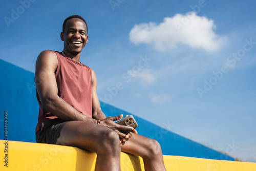 Happy smiling African American man sitting on colourful yellow and blue wall after running and practising sport 