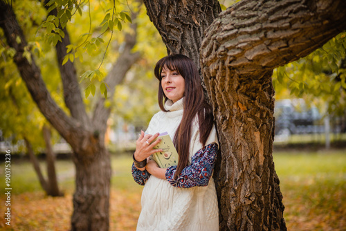 Autumnal thoughtfulness, a moment of reflection: a woman in a white sweater in a park by a tree with a book