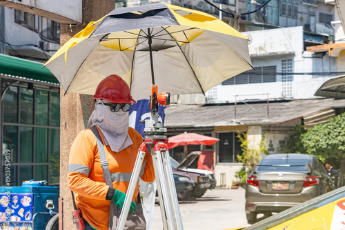 A construction worker using professional surveying tools on street and is sheltered by an umbrella against the strong sun