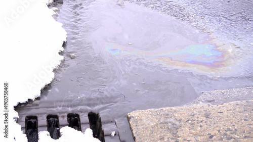 Video of an oil spill mixed with melting snow flowing into a storm drain. Iridescent surface highlights environmental pollution, water contamination, and ecological damage.