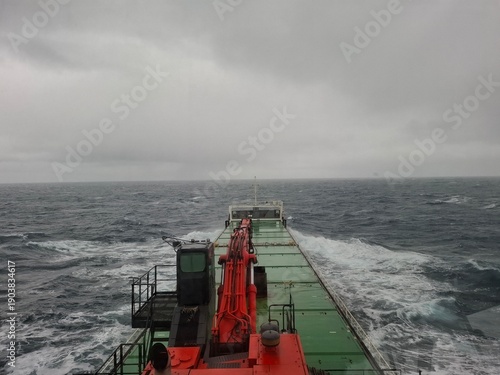 Cargo ship deck with red crane moving through rough sea during stormy weather. Industrial maritime transport and shipping concept with ocean waves and overcast sky.