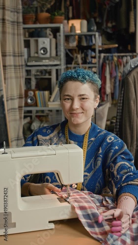 Vertical portrait shot of cheerful young woman with short blue hair and prosthetic arm operating sewing machine while working in modern thrift store