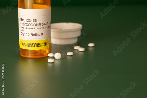 Photography Oxycodone prescription medication in yellow orange plastic bottle with round white tablets out of container on a dark green background