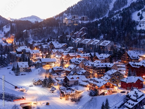 Aerial view of a snow-laden village nestling beneath majestic mountains, lights twinkling like stars against the cold, twilight sky, Claviere, Piedmont, Italy.