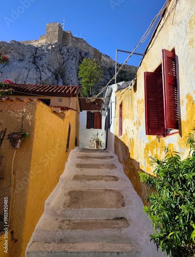 фотографія Photo of a traditional narrow street in Anafiotika neighborhood of Athens, Greece, with white stairs and the Acropolis hill in background