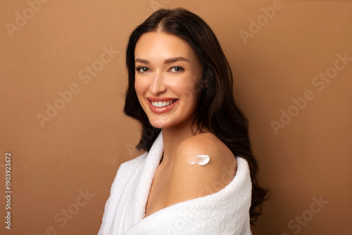 Self-care concept. Young woman applying moisturizing body lotion on shoulder skin, posing over beige studio background and smiling at camera