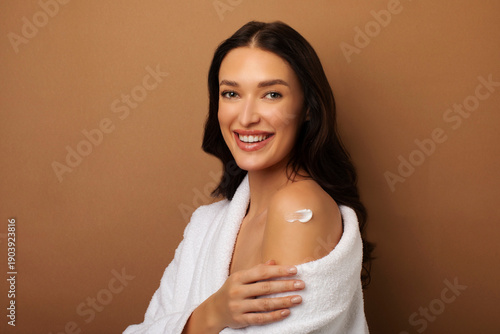 Smiling young woman applying body lotion or cream on shoulder, wearing white bathrobe and posing isolated on beige studio background