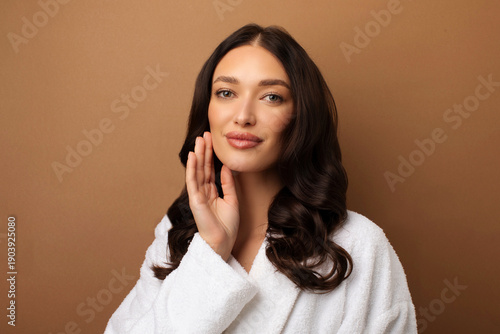 Beauty care. Portrait of young woman with flawless skin touching face and looking at camera, standing on beige studio background