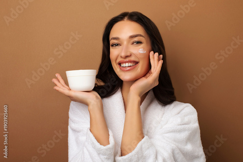 фотографія Cheerful European lady wearing white bathrobe holding jar with nourishing face c