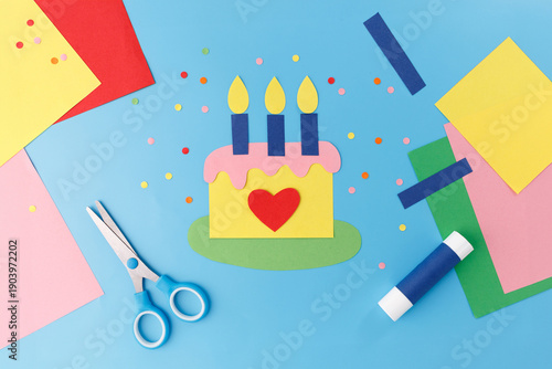 Paper craft birthday cake with candles scissors colorful sheets on blue background. Perfect for creative education, kindergarten workshops, handmade greeting card business, art supplies shop