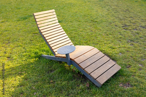 Modern wooden lounge chair with a side table positioned on green grass in a sunny outdoor park setting, designed for relaxation and leisure
