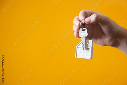 Hand holding silver keys with house shaped keychain on yellow background, concept of real estate and home ownership