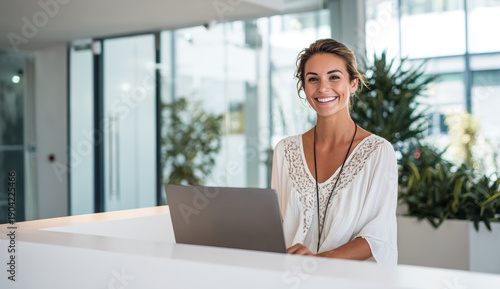 Smiling female receptionist with wavy light brown hair wearing a white embroidered blouse sitting at a reception desk with a laptop in a modern, bright office environment