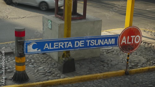A tsunami warning sign next to a stop sign. Both written in spanish. Cars drive by behind it. Located in Miraflores district of Lima, Peru.