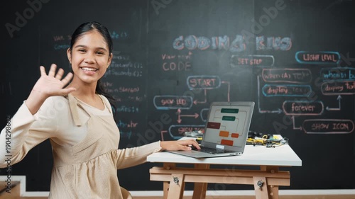 Smart high school girl waving to camera while programming code at blackboard with engineering code or prompt written. Academic student working on laptop or writing program and greeting. Edification