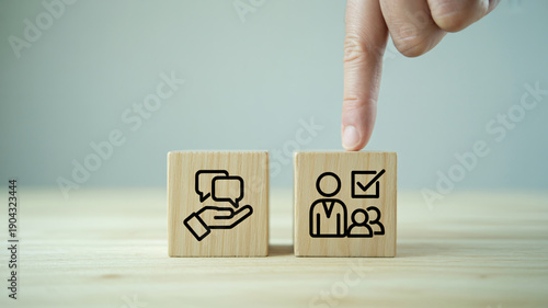 A hand pointing at a wooden block with a human resources icon, next to a communication icon on a light wooden surface.