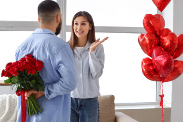 Young man with bouquet of red rose flowers and his surprised girlfriend in living room. Valentine's day celebration