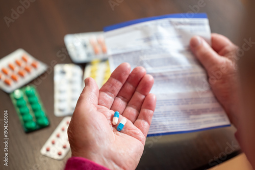 Elderly woman holding medication and the corresponding instruction leaflet. Hands of a pensioner holding two pills in one hand and medication instructions in the other near a table with medicines