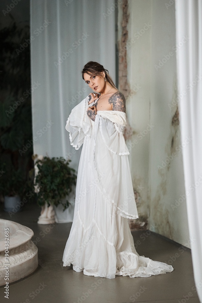 Fototapeta premium Tattooed woman in flowing white dress posing elegantly in a vintage interior setting