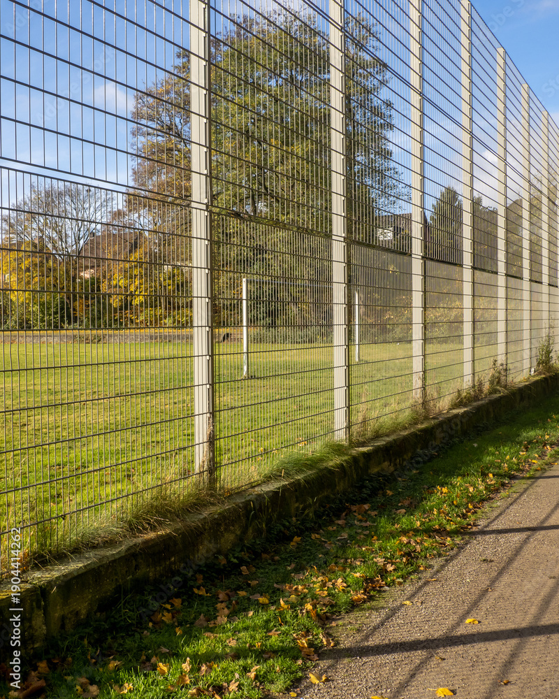 Fototapeta premium Fenced soccer field near a walking path on a clear autumn day with fallen leaves