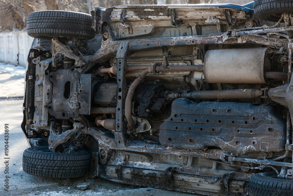 Fototapeta premium A detailed medium shot of the undercarriage and front suspension of an overturned SUV after a road accident. The image shows the complex mechanical components, chassis, and dirty body panels of the ve