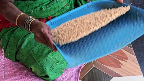 An Indian woman in a green sari is winnowing or sifting grains, likely jowar (sorghum) or rice, using a traditional blue flat basket to separate the grains from the chaff. Its a common rural practice.