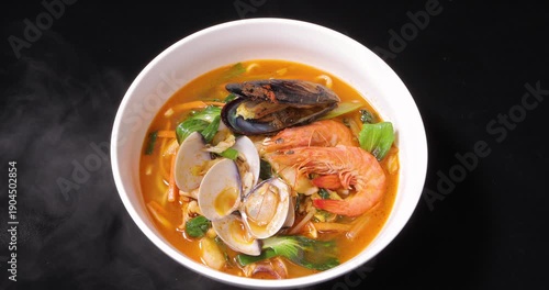 Against a black background, a close-up photo showcases a steaming bowl of Korean spicy seafood soup noodles filled with fresh shrimp, mussels, clams, and vegetables.