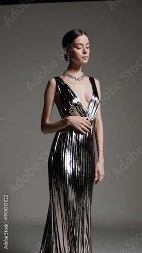 Elegant young woman models a stunning, metallic silver, floor-length evening gown featuring a plunging neckline and intricate back detailing, perfect for a formal gala or red carpet event.