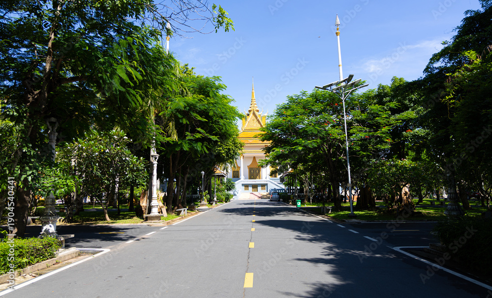 Naklejka premium Majestic Golden Temple Architecture Framed by a Lush Green Tropical Avenue Under a Vibrant Blue Sky.