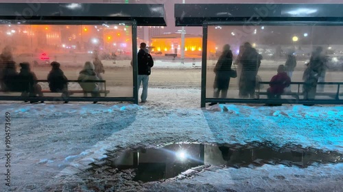 The video opens with a panoramic view of an illuminated bus stop at dusk: the streetlights cast a yellow glow on the snow covering the sidewalk and the roof of the pavilion. Tired passengers in warm 