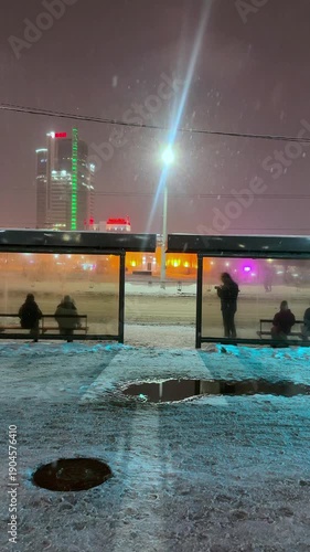 The video opens with a panoramic view of an illuminated bus stop at dusk: the streetlights cast a yellow glow on the snow covering the sidewalk and the roof of the pavilion. Tired passengers in warm 