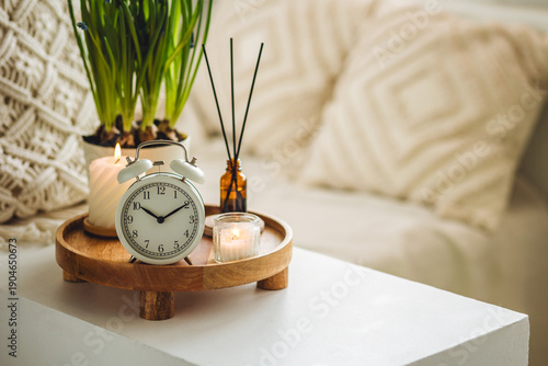 Spring change, Daylight Saving Time concept. White alarm clock, bulbous flowers, candles on the white table. Cozy home interior. Apartment aroma diffuser, aromatherapy. Copy space