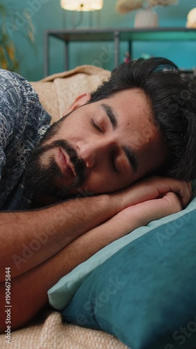 Tired Indian man lying down in bed taking a rest. Carefree young Hindu boy napping, falling asleep on comfortable sofa with pillows at home apartment room. Closed his eyes enjoy daytime nap alone