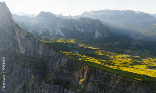 Wide aerial view of Seceda alpine plateau with green meadows and steep limestone cliffs. Summer light reveals the scale and beauty of the Dolomites landscape