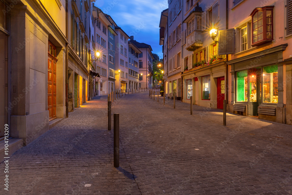 Obraz premium Early morning view of an empty cobblestone street in Zurich old town, lined with traditional buildings and softly glowing streetlights