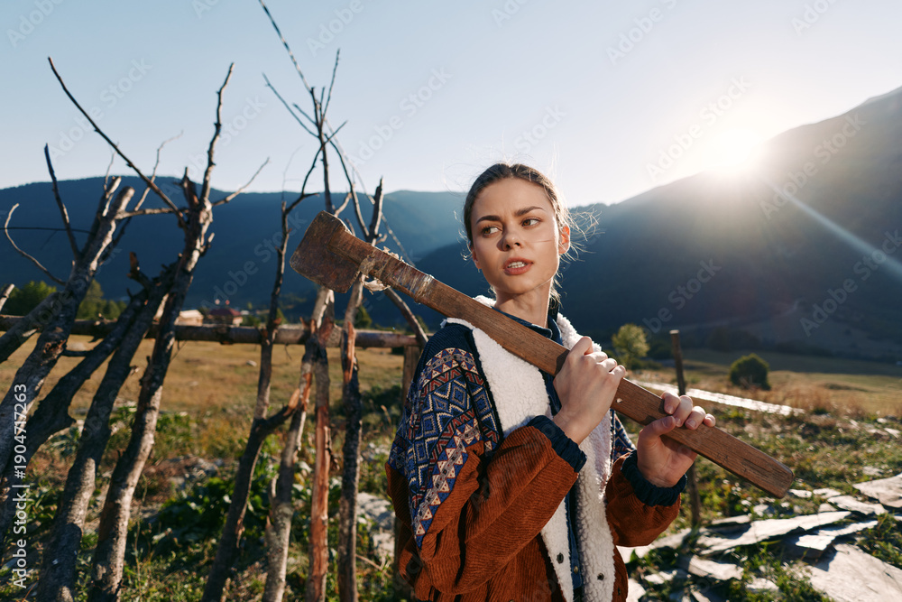 Fototapeta premium Woman carrying an axe over her shoulder in rural countryside with mountains and bright sunlight, farming fence and fields, outdoor portrait of a young worker in nature.
