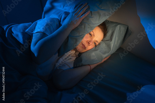Middle-aged woman lying in bed covering ears with pillows due to noisy neighbors, stressed female suffering from insomnia and loud music at night