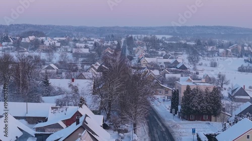 4K aerial drone tracking right over Semeliškės, Lithuania, revealing snowy main street and rooftops at winter sunset, small town cinematic cityscape footage